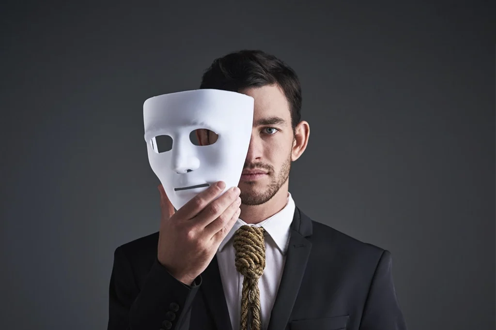 Hombre sosteniendo una máscara blanca frente a su rostro, simbolizando el síndrome del impostor y la falta de merecimiento tratada con hipnoterapia clínica.